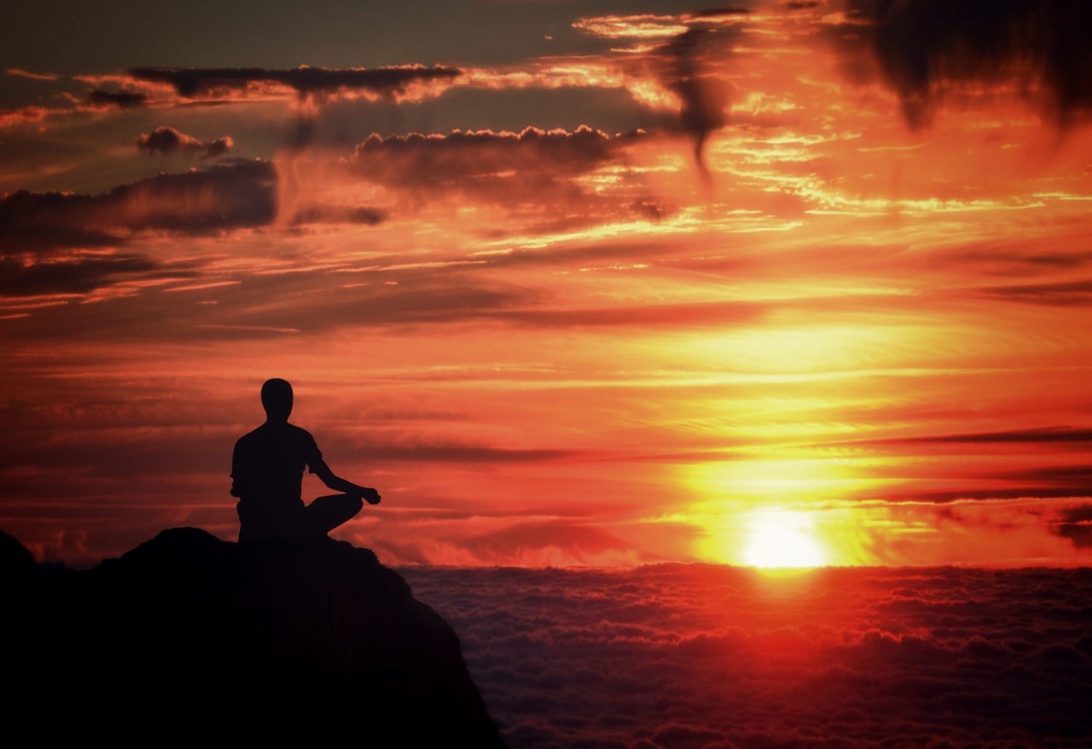 En silhuett av en person som sitter i meditationsställning på en sten eller bergstopp, mot en dramatisk orange och röd solnedgångshimmel. Kompositionen skapar en kraftfull visuell representation av frid, ensamhet och andlig koppling under meditationsövning.