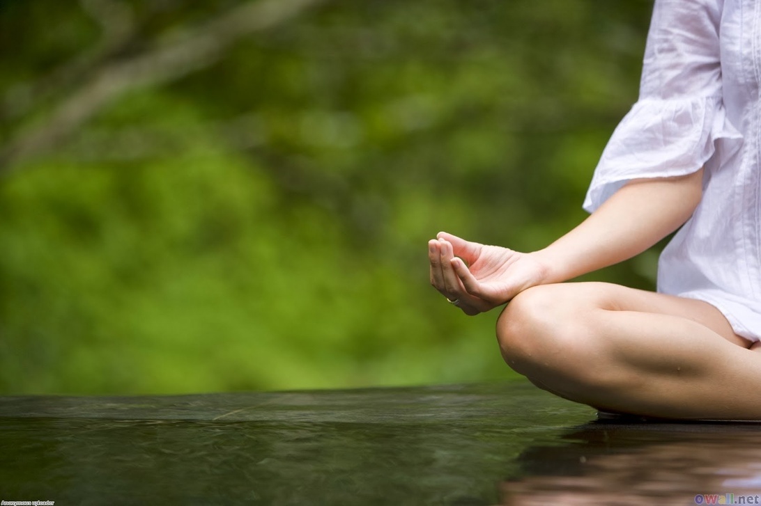 En delvis vy av en person i vita kläder som sitter i meditationsställning nära vatten. Bilden fokuserar på handen i en meditationsmudra och korslagda ben. Den naturliga gröna bakgrunden och vattnet skapar en fridfull meditationsmiljö och betonar kopplingen till naturen.
